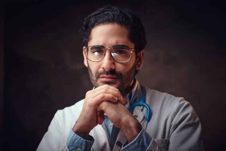 Pensive physician with a stethoscope, hands clasped under his chin, against a dark background