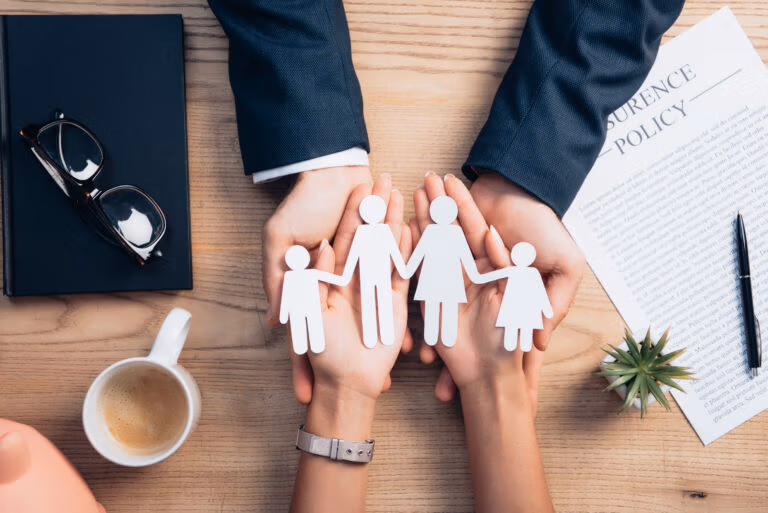 Hands holding a paper-cutout family over a desk with coffee and an insurance policy document