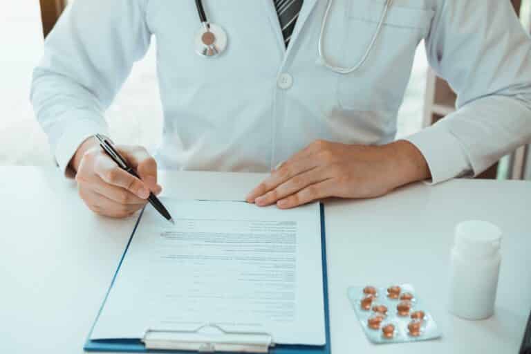 Doctor in a white coat filling out a form on a clipboard, with medication on the desk