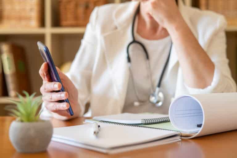 Doctor in a white coat with a stethoscope using a smartphone at a desk with notebooks