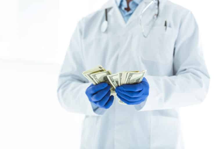Doctor in a white coat and blue gloves counting U.S. dollar bills