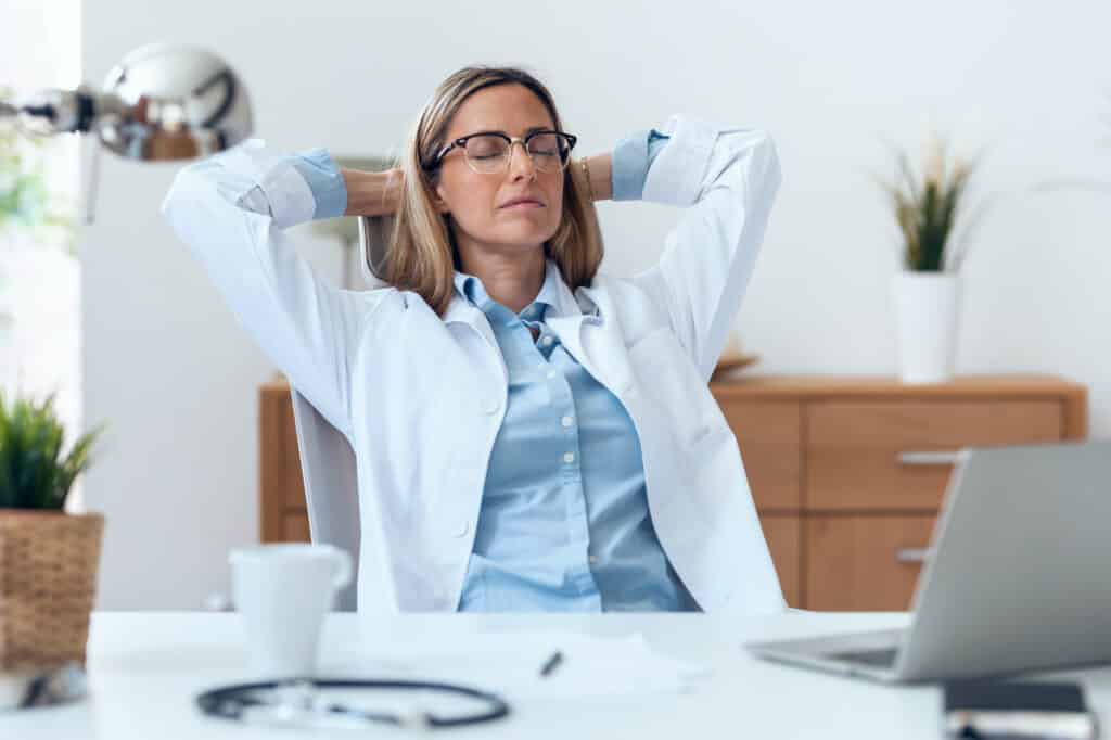 Doctor in a white coat leaning back in her chair, stretching at her desk