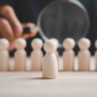 A single wooden figurine stands out from a row of similar figures, highlighted by a magnifying glass.