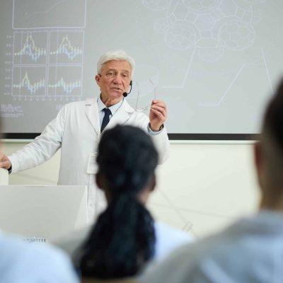 Senior scientist presenting research findings to attentive students in modern classroom environment with graphs displayed on the screen while making important points