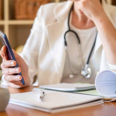 Healthcare professional wearing a stethoscope and white coat, using a smartphone for telemedicine or remote patient care, working at a desk in a medical office environment